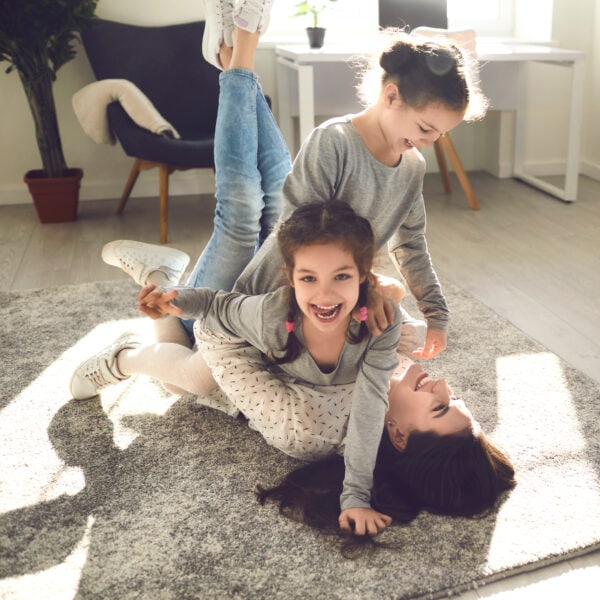 Family enjoying quality time and having fun at home. Young mom and little kids playing on carpet backlit with sunlight. Happy mother and cute carefree children fooling around and laughing on floor rug